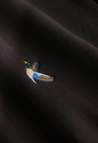 Czarna tkanina z szczegółowym haftowanym wizerunkiem kaczki z zielonymi, niebieskimi i brązowymi akcentami, osadzonym na teksturowanym tle.