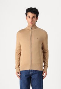 Pull en maille beige zippé avec un col montant, des poignets et un ourlet côtelés. Possède un petit logo sur la poitrine. Assorti à un jean en denim bleu.