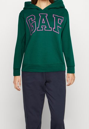 Kobieta ubrana w zieloną bluzę z kapturem z dużymi różowymi literami „GAP” na piersi oraz ciemne spodnie, stojąca na białym tle.