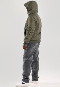 Olivově zelená bunda s kapucí, elastickými manžetami, bočními kapsami a logem na rukávu, doplněná šedými džínami a bílými teniskami.