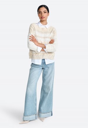 Pull en tricot beige avec des rayures texturées, superposé à une chemise blanche à col. Jean large bleu clair avec des ourlets retroussés et des chaussures de couleur claire.