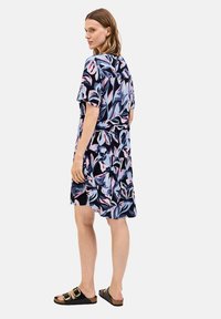 Blumengemustertes Kleid aus blauem, pinkem und schwarzem Stoff mit kurzen Ärmeln, tailliertem Schnitt und gestuften Saum. Getragen mit schwarzen Sandalen.