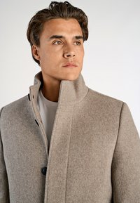Ung mand med kort brunt hår iført en beige uldfrakke over en hvid skjorte, der kigger lidt til siden med en neutral udtryk.