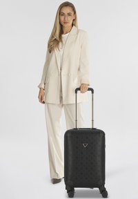 Maleta rígida negra con patrón en relieve, asa telescópica y cuatro ruedas, acompañada de una americana y pantalones de color claro.