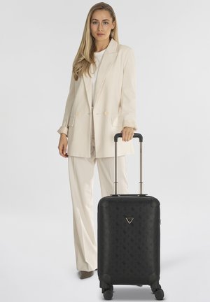 Valigia rigida nera con motivo in rilievo, manico telescopico e quattro ruote, abbinata a un blazer e pantaloni chiari.