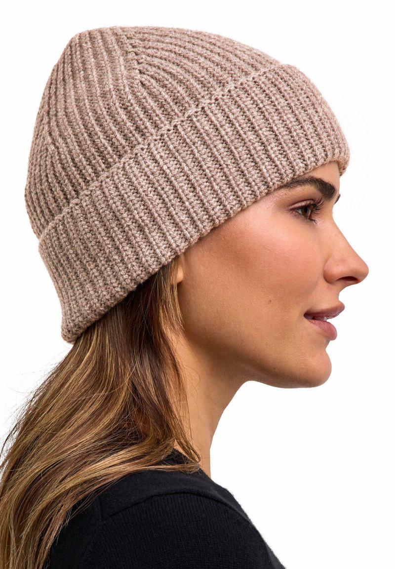 Woman with light brown hair wearing a beige ribbed knit beanie and a black top, shown in profile on a white background.