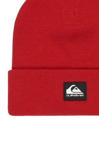 Red knitted beanie with a folded cuff, featuring a black rectangular logo patch with white text and graphic design.