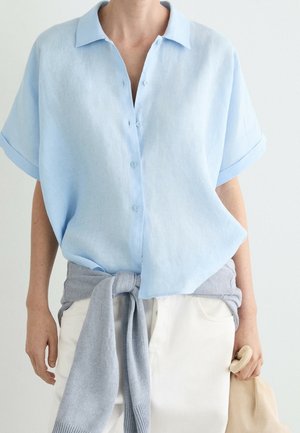 Personne portant une chemise bleu clair à manches courtes boutonnée, avec un pull gris noué autour de la taille et tenant un sac beige.