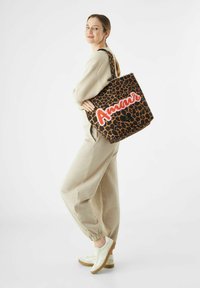 Vrouw in een beige outfit en witte sneakers, met een grote tassen in luipaardprint met rode "Amour" tekst op haar schouder.