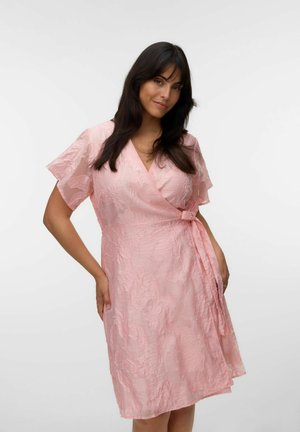 Kvinde med langt mørkt hår iført en lyserød tekstureret omslagskjole med flagrende ærmer, poserer foran en ensfarvet hvid baggrund.