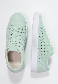 Mátově zelené perforované tenisky s velurovým svrškem, bílou gumovou podrážkou a šněrováním. Hladká textura a kulatý tvar špičky.