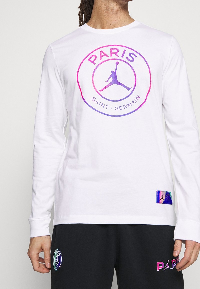 Vit långärmad tröja med en cirkulär grafiklogo med "PARIS" och "SAINT-GERMAIN" i lila och rosa. Inkluderar en etikett med holografiska detaljer.