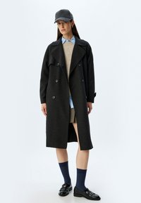 Zwart wollen jasje met een double-breasted ontwerp, grote kraag en voorzakken. Gecombineerd met een lichtblauw shirt en een beige sweater. Zwarte pet, kniekousen en loafers.