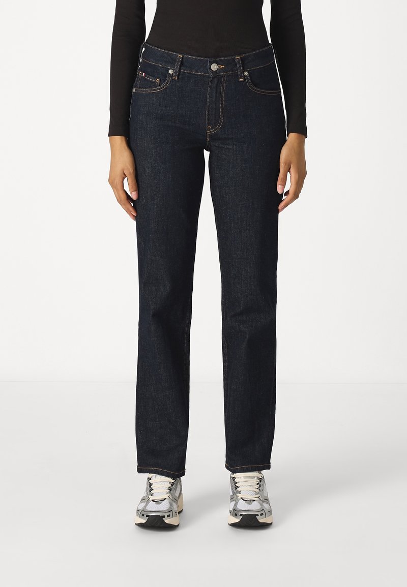 Personne portant un jean droit bleu foncé, un haut noir à manches longues et des baskets argentées et blanches, debout sur un fond blanc.