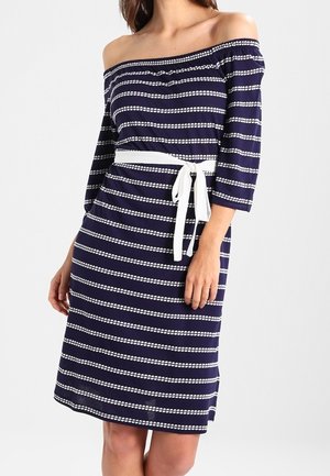 Femme portant une robe bleu marine épaules dénudées avec des rayures blanches horizontales en pointillés et une ceinture blanche nouée à la taille.