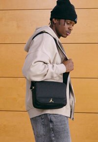 Sort Jordan skulderveske med gullfarget logo, båret på tvers over en lys beige hettegenser, kombinert med grå jeans mot en trebakgrunn.