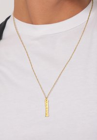 Goldfarbener Halsreifen mit einem rechteckigen Anhänger, der das Wort "FRIEDEN" eingraviert hat. Die Kette ist zart mit einem gedrehten Design.