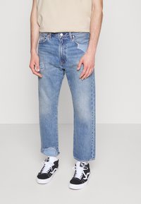 Ljusblå denimjeans med avslappnad passform, som har en slitna lapp på vänster lår och fransig fåll, kombinerat med svarta sneakers.