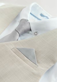 Ljusbeige kavaj med matchande sidensjalkficka över en vit skjorta och ljusgrå slips med subtilt mönster.