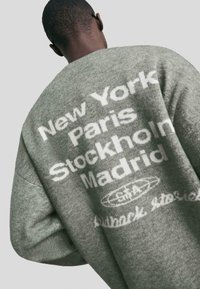 Eine Person trägt einen grauen Pullover mit weißem Text, der die Städte New York, Paris, Stockholm und Madrid auf dem Rücken auflistet, vor einem neutralen Hintergrund.