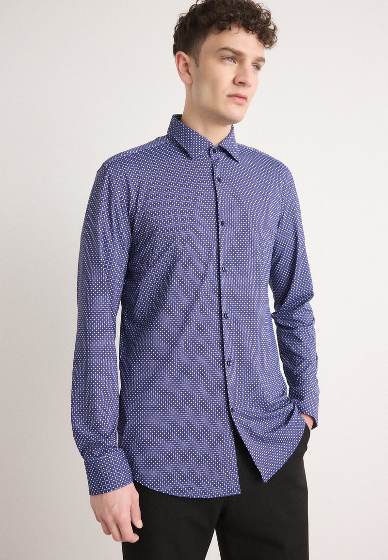 Camisa azul marino con un pequeño patrón de puntos blancos, cierre de botones, mangas largas y cuello puntiagudo. Textura suave y ajuste entallado.