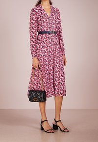 Šaty s květinovým potiskem, dlouhým rukávem, výstřihem do V, páskem v pase a splývavou sukní; doplněné černou prošívanou kabelkou a sandály na blokovém podpatku.