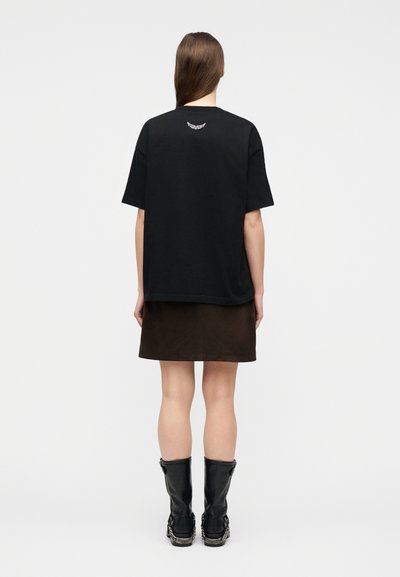 Γυναίκα με μακριά μαλλιά που φοράει μαύρο φαρδύ T-shirt με μικρό λευκό έμβλημα στο πάνω μέρος της πλάτης, καφέ φούστα και μαύρες μπότες, όρθια.