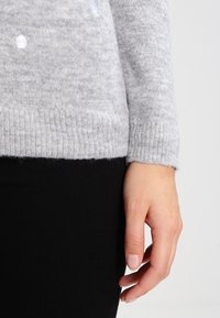 Ljusgrå stickad tröja med ribbad kant och subtil textur. Svarta byxor är delvis synliga. Handen vilar p å sidan.