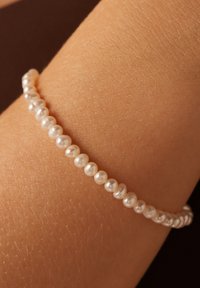 Parelarmband met kleine, ronde witte parels, met een licht onregelmatig oppervlak, aan elkaar geregen. Tegen een текстурированным huidoppervlak.