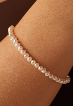 Bracelet en perles avec de petites perles blanches rondes, surface légèrement irrégulière, enfilées ensemble. Placé sur une surface de peau texturée.