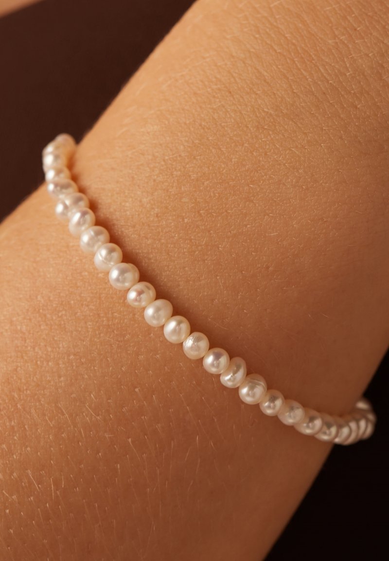 Parelarmband met kleine, ronde witte parels, met een licht onregelmatig oppervlak, aan elkaar geregen. Tegen een текстурированным huidoppervlak.