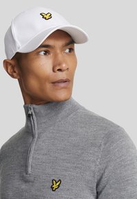 Weißer Baseballcap mit gelbem besticktem Logo, kombiniert mit einem grauen Pullover mit Reißverschlusskragen und passendem Logo auf der Brust.