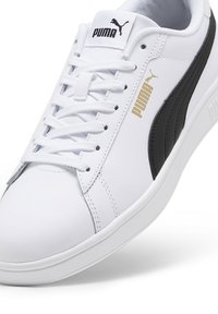 Baskets synthétiques blanches avec accents noirs, dotées d'une tige texturée, de lacets plats et d'un logo Puma doré sur le côté et la languette.