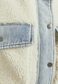 Jeansjacka med kropp i krämfärgad, fluffig fleece och blå jeansdetaljer. Inkluderar silverknappar och en framficka.