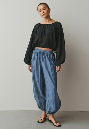 Młoda kobieta ubrana w luźną czarną bluzkę z długim rękawem, niebieskie spodnie ze ściągaczami i czarne sandały na obcasie, stojąca na tle jednolitej ściany.