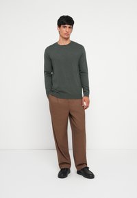 Mörkgrön långärmad tröja i kombination med lösa bruna byxor och svarta sneakers; mjuk textur och subtila sömdetaljer.