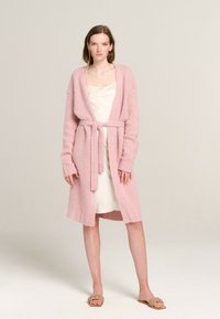 Langer, rosa gestrickter Cardigan mit offenem Front, tailliertem Gürtel und gerippten Bündchen. Getragen über einem cremefarbenen Kleid und kombiniert mit tanfarbenen geflochtenen Sandalen.