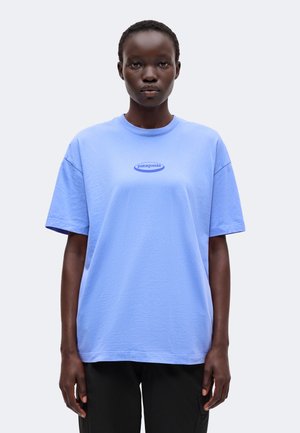 Personne aux cheveux courts portant un t-shirt Patagonia bleu clair et un pantalon noir, debout devant un fond clair uni.