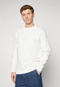 Fehér pamut pulóver, kerek nyakkal, bordás mandzsettákkal és szegéllyel, valamint a melléhez hímzett logóval, kék farmernadrággal párosítva.