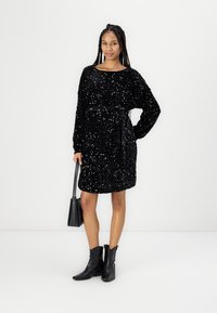 Vestido preto de lantejoulas com mangas longas, corte descontraído, cinto na cintura. Usado com botas pretas até ao tornozelo e a carregar uma pequena mala preta.