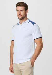 Homme portant un polo blanc avec des accents bleus marine sur les épaules et un logo « Hackett », assorti à un pantalon beige, debout devant un fond uni.