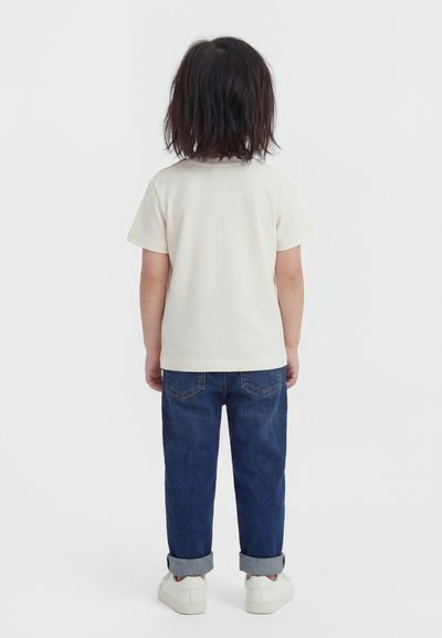 Παιδί με σκούρα μαλλιά μέχρι τους ώμους, που φοράει λευκό κοντομάνικο μπλουζάκι, μπλε τζιν με γυρισμένα ρεβέρ και λευκά αθλητικά παπούτσια, στέκεται γυρισμένο προς τα πίσω.