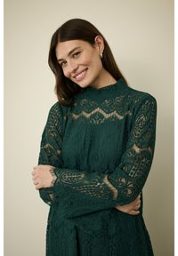 Zelený krajkový šaty v barvě smaragdu s dlouhými rukávy a vysokým výstřihem, které mají složité vzory a průhledné detaily. Modelka pózuje se zkříženýma rukama.