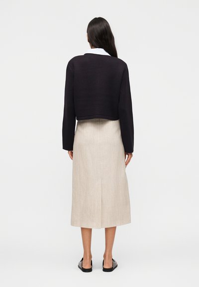 Γυναίκα με μακριά σκούρα μαλλιά φοράει μαύρο cropped πουλόβερ, μπεζ midi φούστα με σκίσιμο πίσω, μαύρα slip-on παπούτσια, στέκεται στραμμένη προς λευκό φόντο.