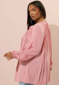 Blusa leggera rosa chiaro con maniche lunghe, dettaglio in pizzo lungo la parte frontale e le maniche, e design con colletto arricciato.