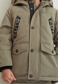 Khaki padded jacket with a hood, featuring black buttons, two front pockets, and "FUTR" printed in green on either side.