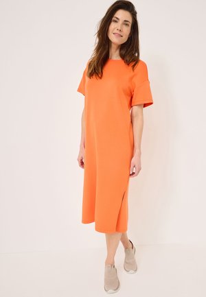 Kurzes orangefarbenes Kleid mit lockerer Passform, kurzen Ärmeln, seitlichem Schlitz und glattem Stoff. Kombiniert mit beigen Slippern.