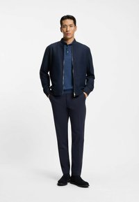 Granatowa kurtka bomberka z zapięciem na zamek, pod spodem niebieska koszulka polo oraz dopasowane granatowe spodnie. Modelka ma na sobie czarne botki za kostkę. Prostota i nowoczesny design.