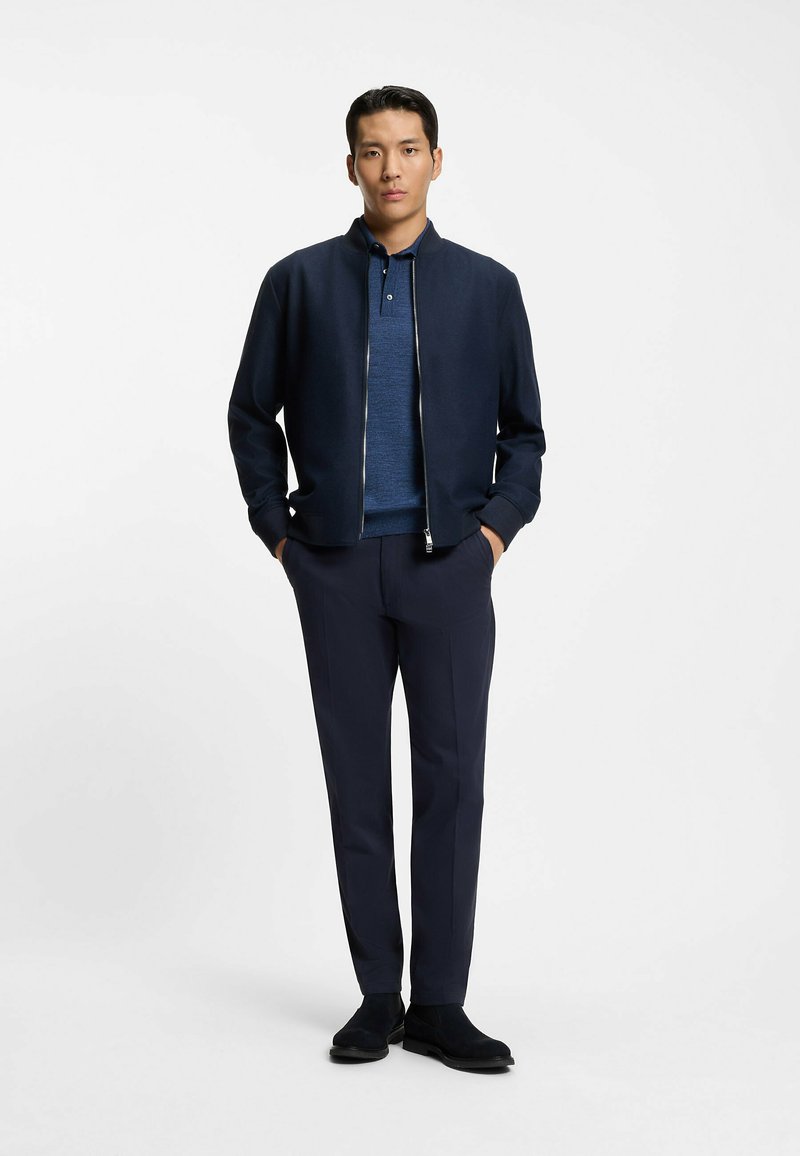 Granatowa kurtka bomberka z zapięciem na zamek, pod spodem niebieska koszulka polo oraz dopasowane granatowe spodnie. Modelka ma na sobie czarne botki za kostkę. Prostota i nowoczesny design.