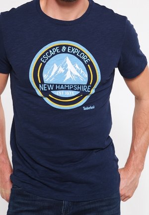 Homme portant un t-shirt bleu marine avec un motif de montagne et le texte "Escape & Explore New Hampshire Est 1973" ainsi que le logo Timberland.